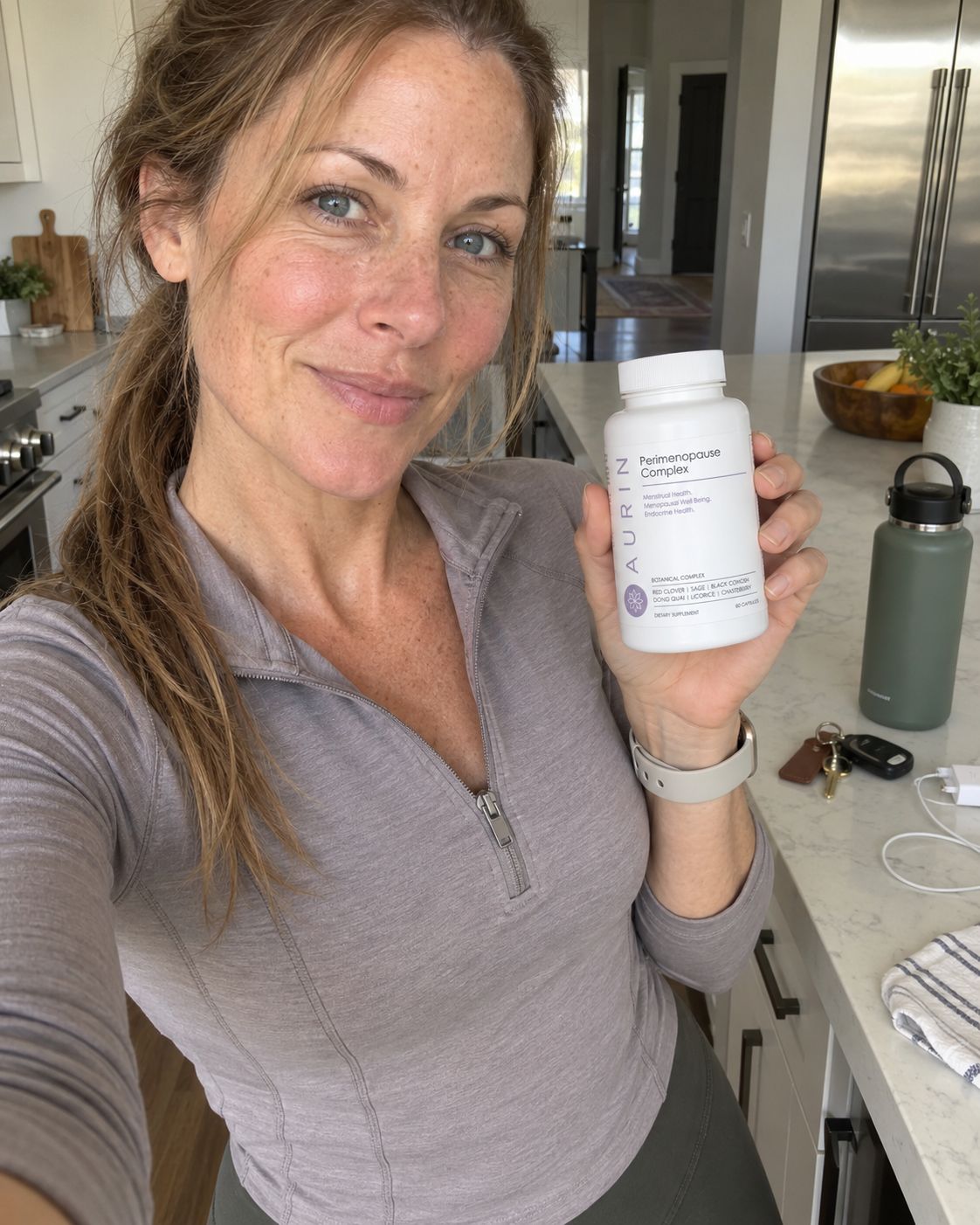 A smiling woman in her kitchen holding a bottle of AURIN Perimenopause Complex